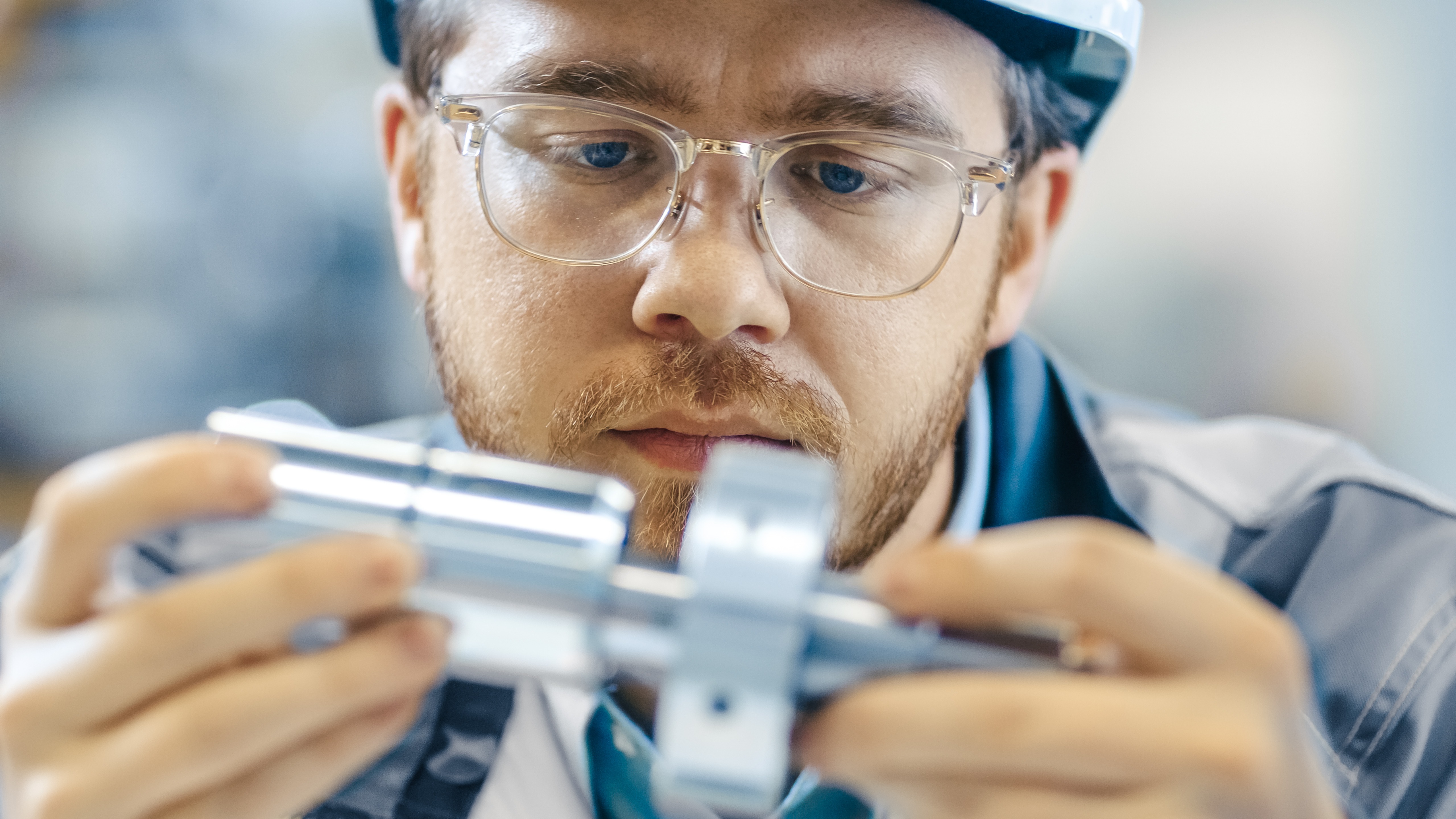 An engineer with a full beard and glasses carefully examining a workpiece