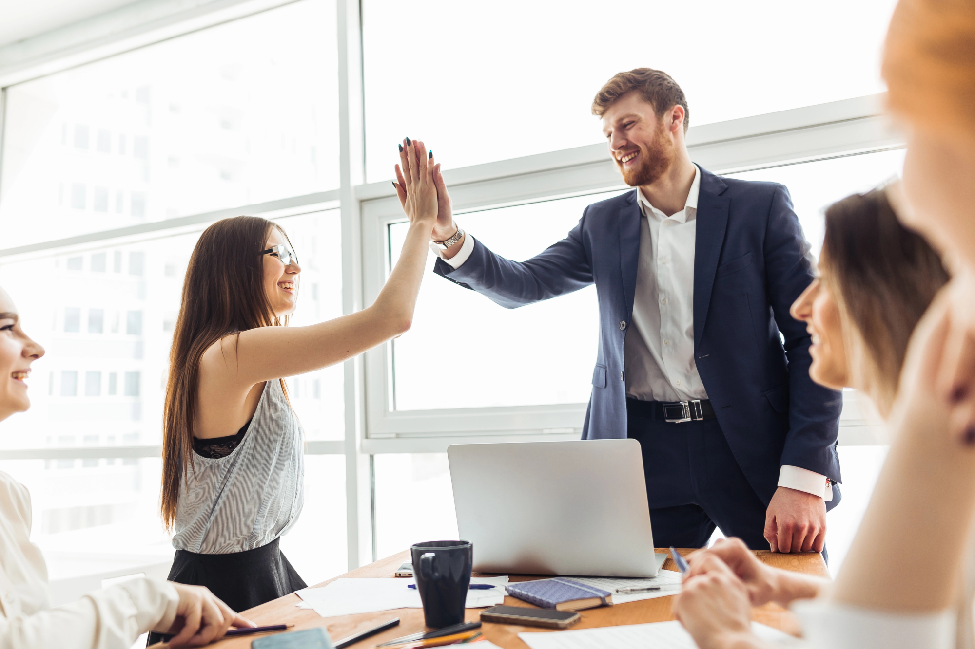 Two cheerful young businessmen high-fiving each other while their colleagues look at them and smile.