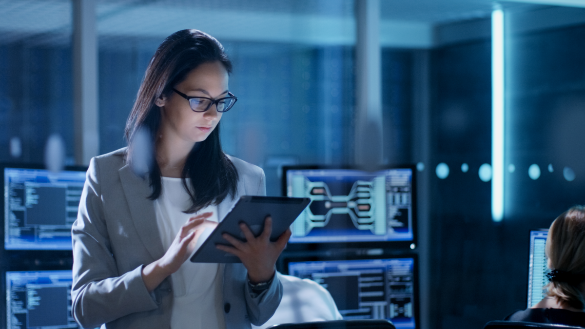 Young Female Employee Wearing Glasses Uses Tablet in System Control Center. In the Background Her Co