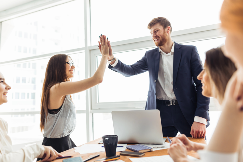 Two cheerful young businessmen high-fiving each other while their colleagues look at them and smile.